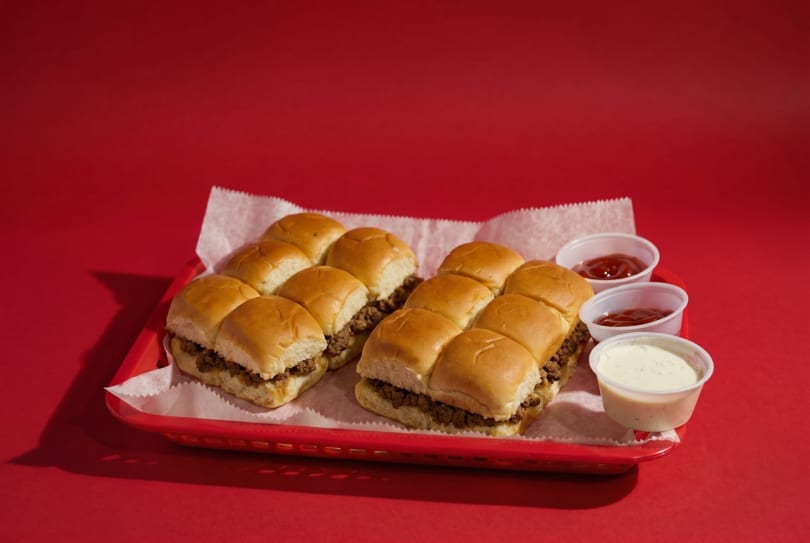 Two rows of sliders with ground meat on a red tray with ketchup and ranch dipping sauces.