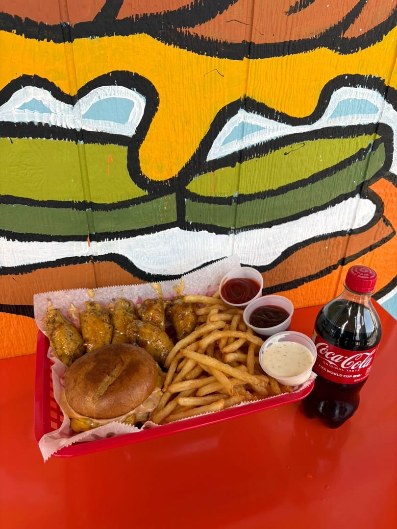 A red basket with a burger, fries, chicken wings, dipping sauces, and a Coca-Cola bottle.