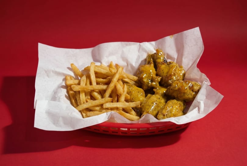 A red basket holds crispy french fries and mustard-glazed chicken wings on white paper.